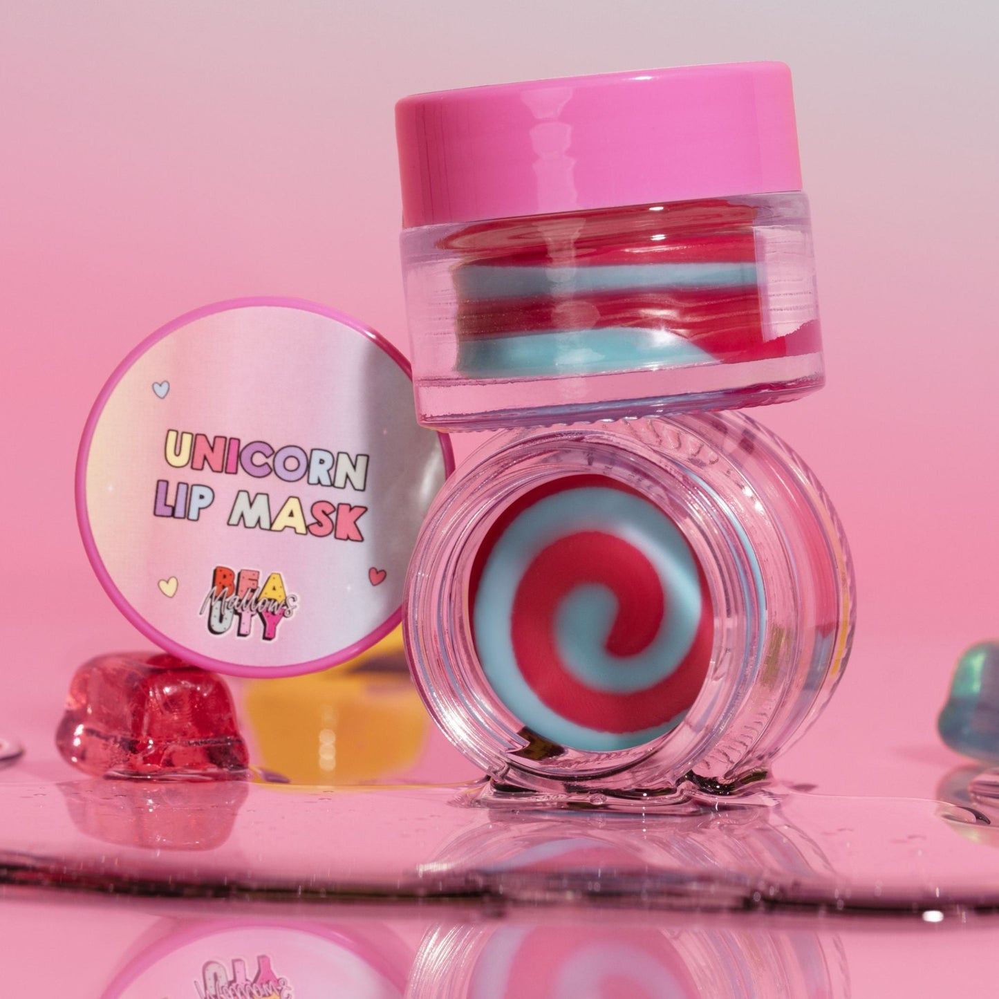 A colorful lip mask product with a unicorn design, presented in a clear container with a pink lid, alongside a spoon and a swatch showing the product applied to lips.