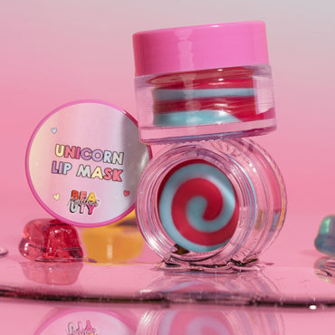 A colorful lip mask product with a unicorn design, presented in a clear container with a pink lid, alongside a spoon and a swatch showing the product applied to lips.