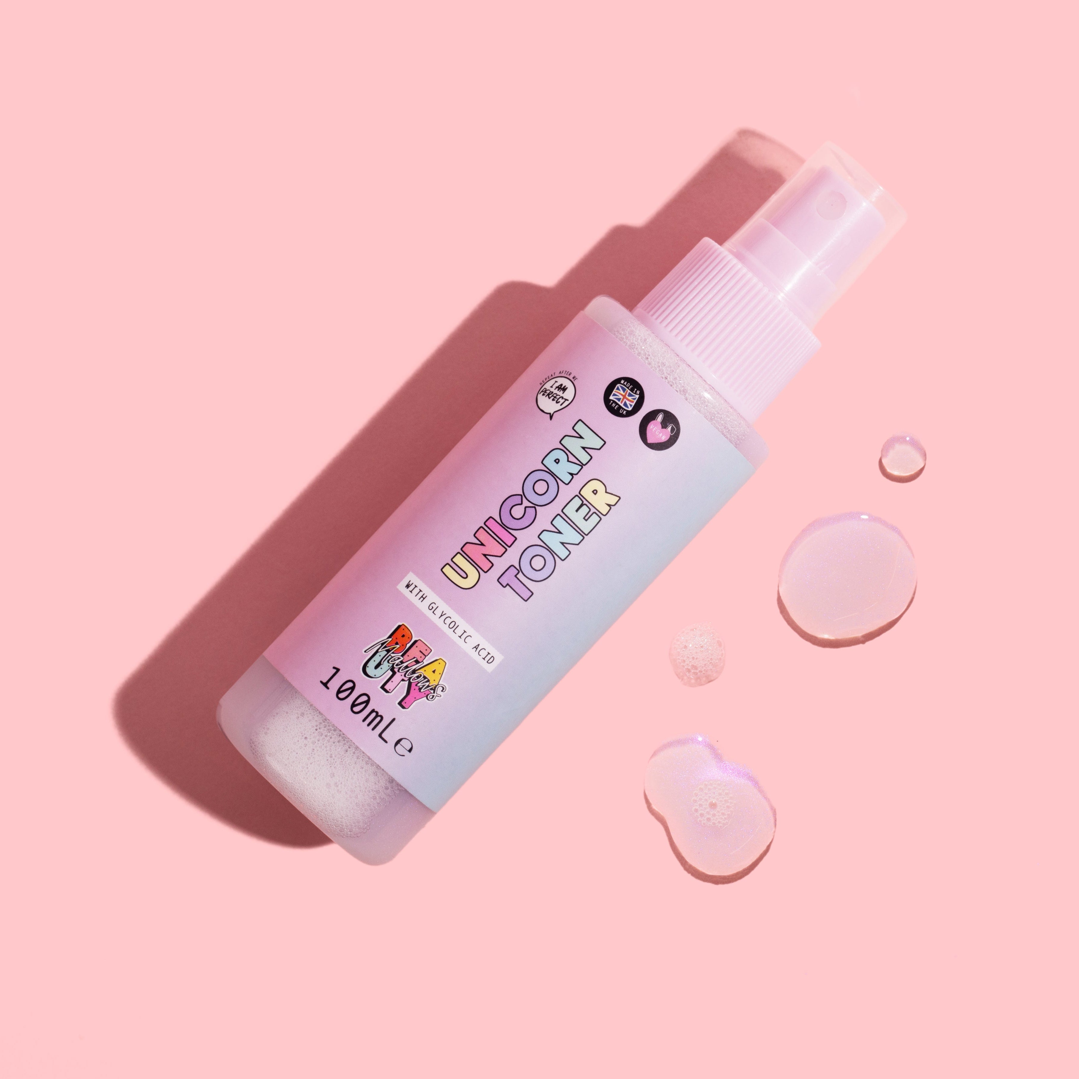A spray bottle labeled 'Unicorn Toner with Glycolic Acid' against a pink background, with a few droplets of liquid visible in front of the bottle.